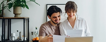 Couple souriant, assis devant un ordinateur et tenant en main des papiers